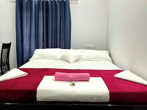 A hotel room with a bed, a chair, a window with blue curtains, and a wall socket.