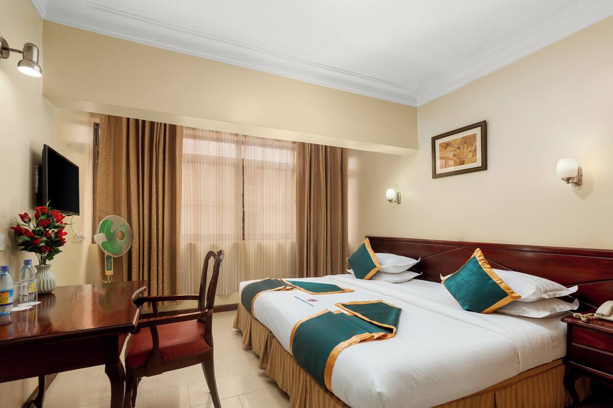 Habitación de hotel con cama, escritorio, silla, televisor, ventilador y ventana con cortinas.