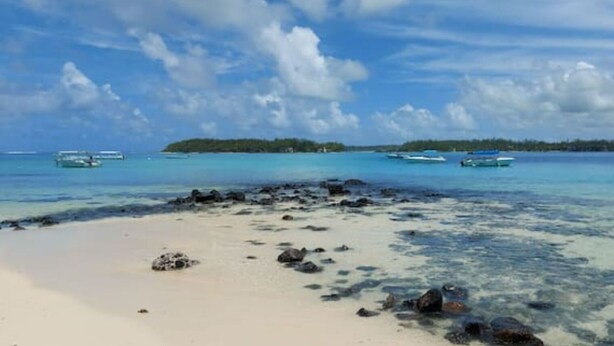 Ein Strand mit klarem türkisblauem Wasser, verstreuten Felsen und Booten in der Ferne.