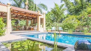 A swimming pool with a small pond in the foreground, surrounded by lush greenery and a covered seating area with chairs and tables.