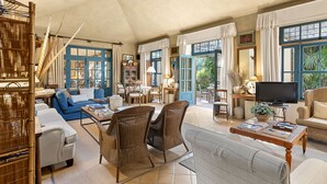 A spacious living room with a blue sofa, wicker chairs, a coffee table with books, a television, and large windows with curtains.