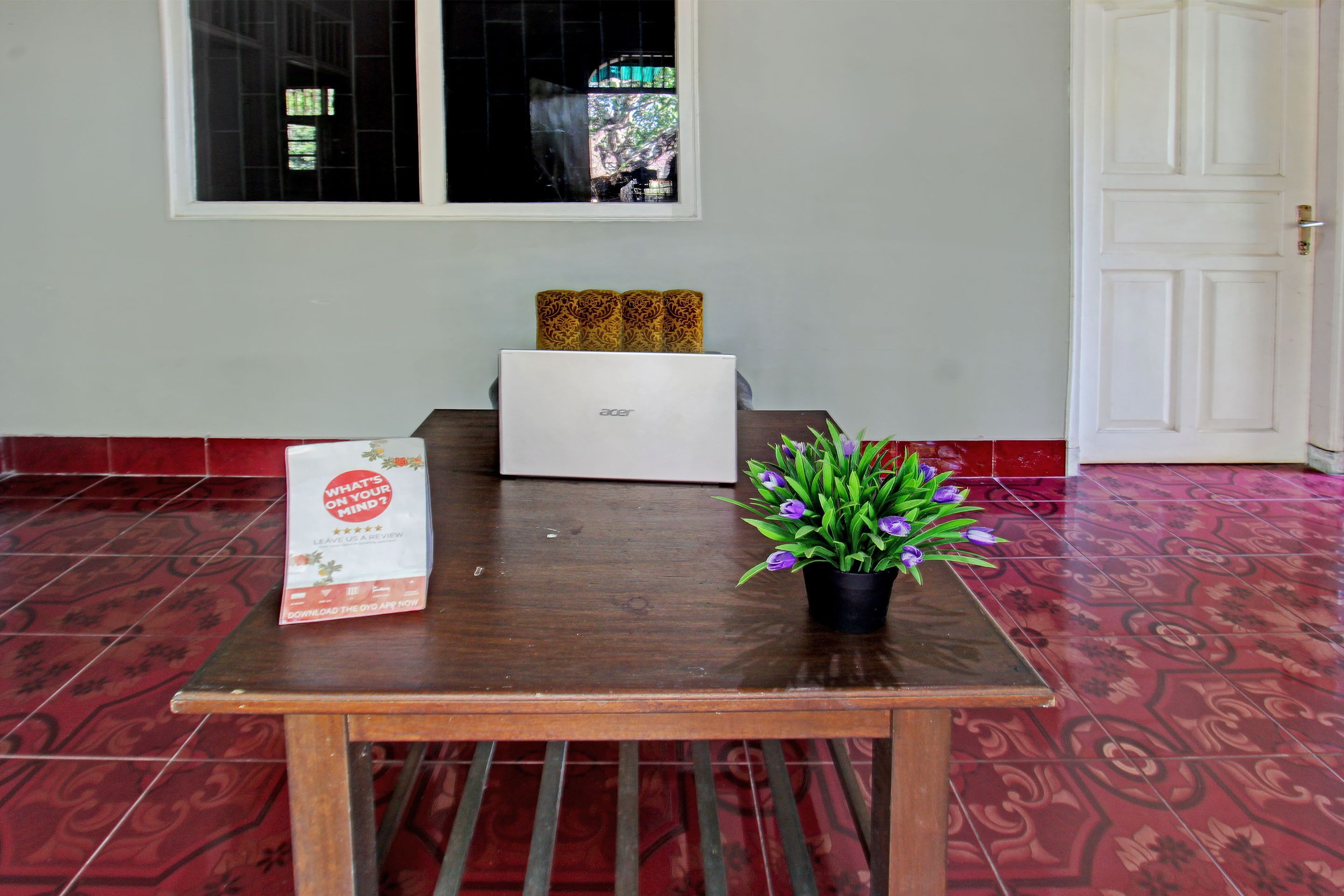 Uma mesa de madeira com um laptop, um vaso de planta e uma sacola promocional em um quarto com piso estampado em vermelho e uma porta branca.