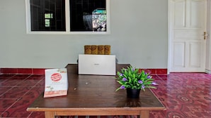 A wooden table with a laptop, a potted plant, and a promotional bag in a room with patterned red flooring and a white door. - SPOT ON 92973 Madani Syariah Guesthouse (Praya)