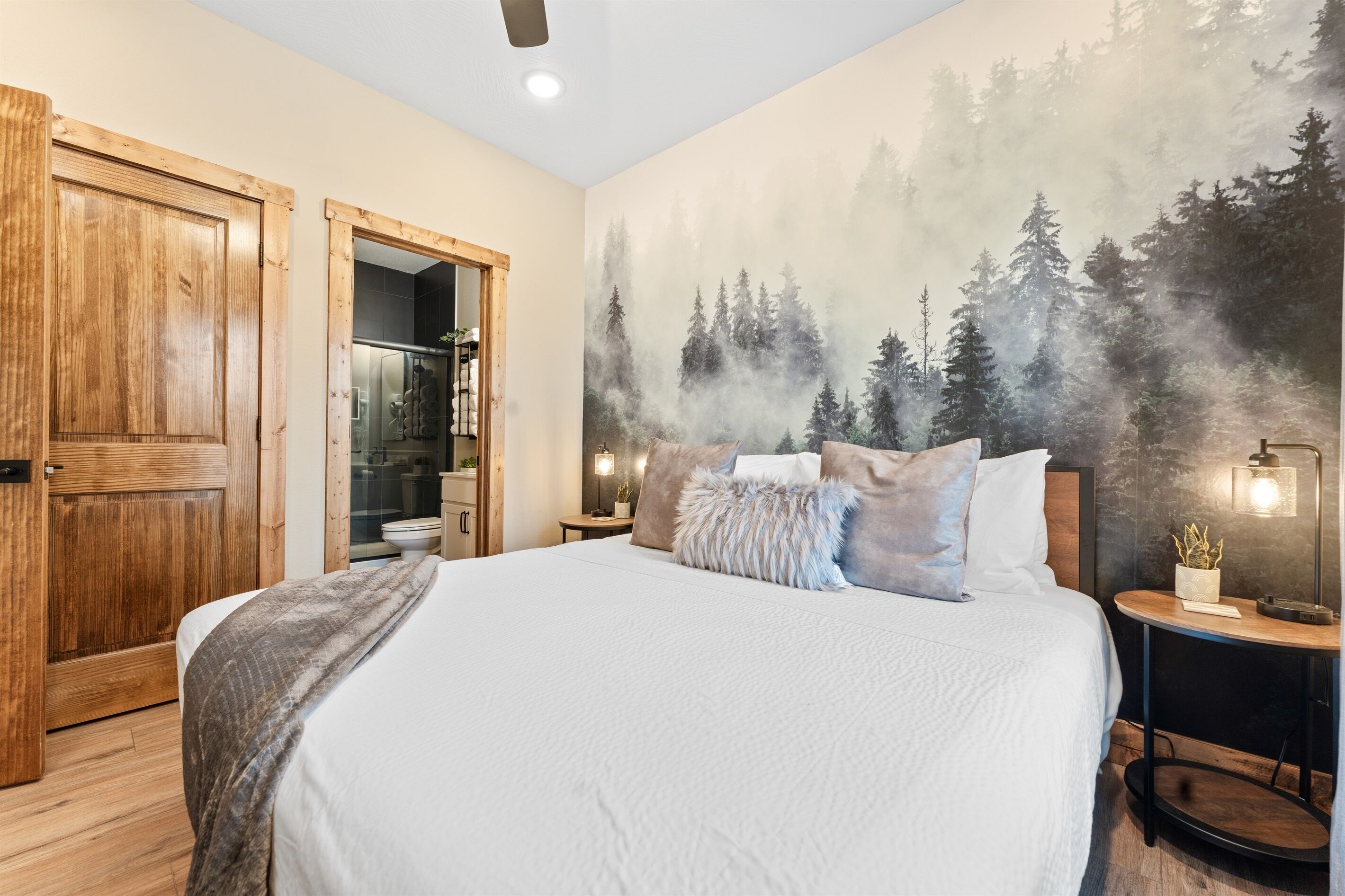 A bedroom with a large bed, a wooden door, a bathroom with a glass shower, and a forest mural on the wall.