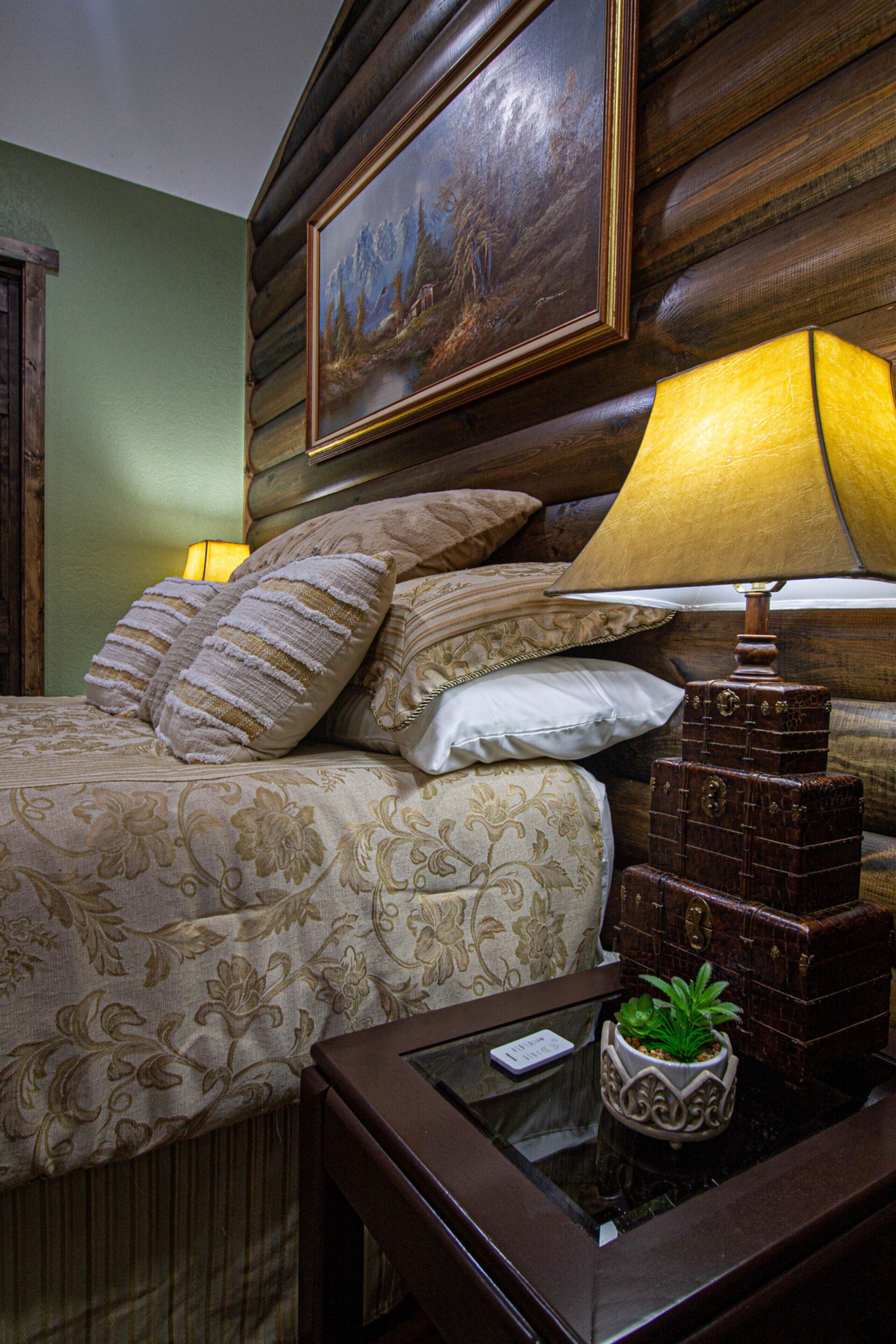 A neatly made bed with a floral bedspread, a wooden headboard, and a painting above it. There is a bedside table with a lamp and a small potted plant.