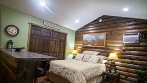 A rustic log cabin-style bedroom with a wooden bed, two bedside lamps, a wooden dresser, and a framed picture on the wall.