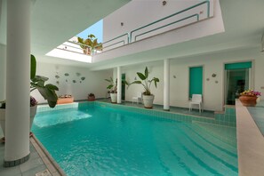 En inomhuspool med kristallklart blått vatten, omgiven av vita väggar och pelare, och dekorerad med krukväxter.