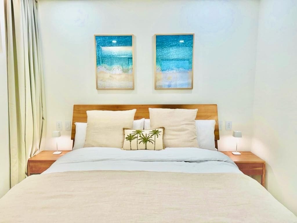 A neatly made bed with a wooden headboard, two wall-mounted paintings of a beach scene, and two bedside tables with lamps.