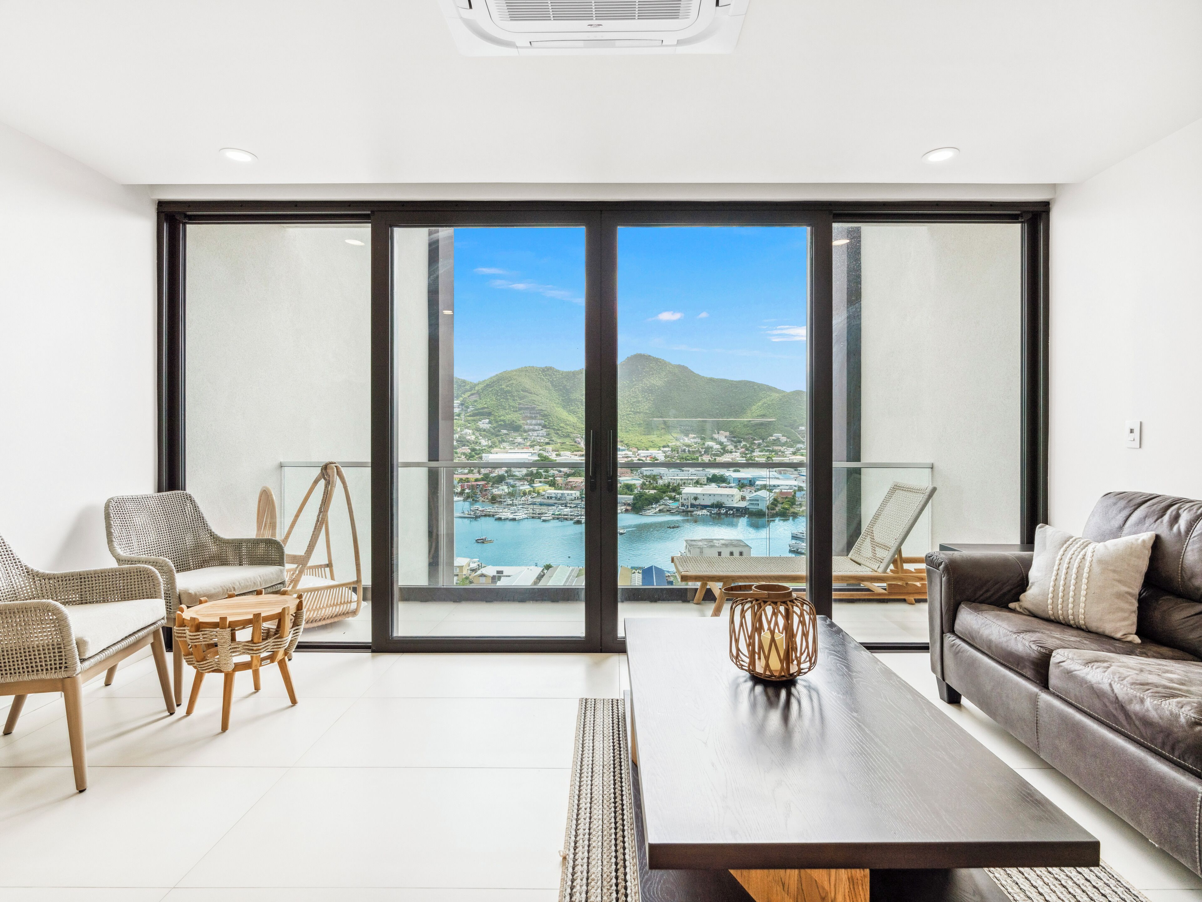 Un salon moderne doté d’une grande porte-fenêtre coulissante qui donne sur un balcon offrant une vue sur une marina et des collines.