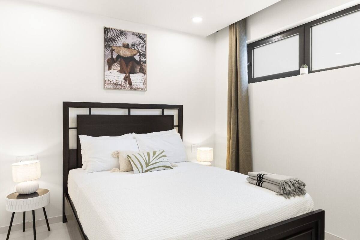 Ein ordentlich bezogenes Bett mit weißen Leinen und einem dunklen Kopfteil in einem modernen Zimmer, mit einem einzelnen Wandbild und einem kleinen Nachttisch mit Lampe.