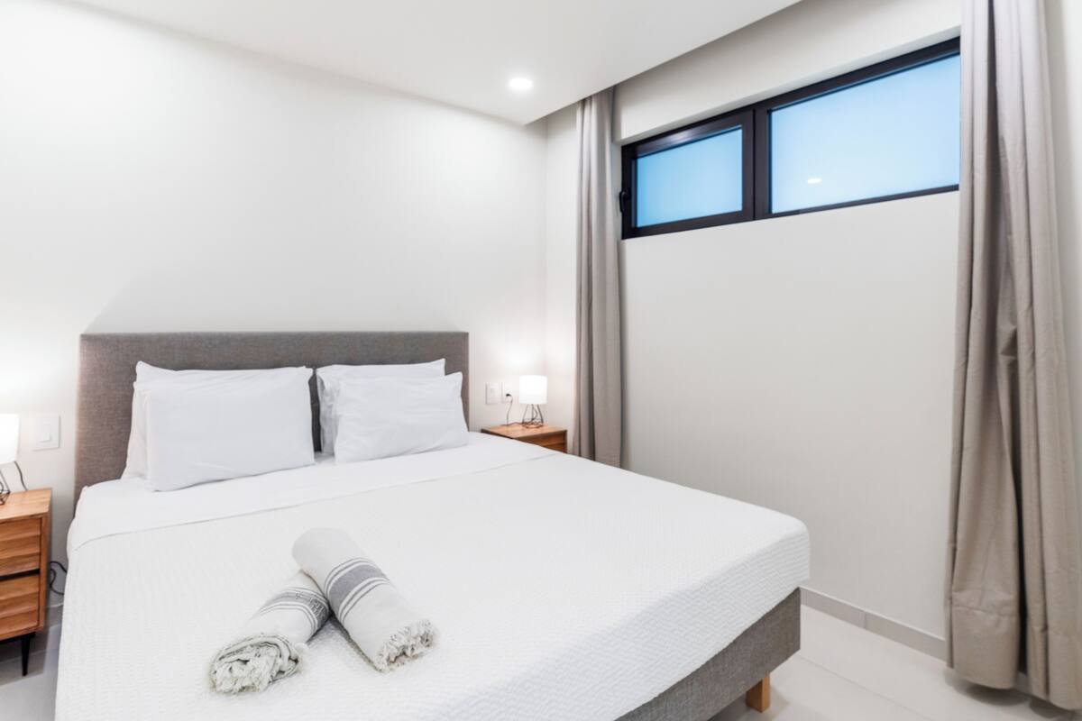 Ein modernes Hotelzimmer mit einem großen Bett, einem Nachttisch, einem Fenster mit Vorhängen und Blick auf den Himmel.