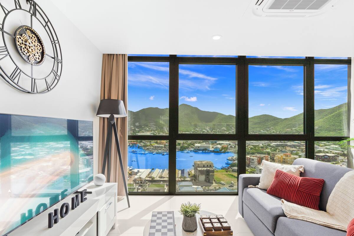 Ein modernes Wohnzimmer mit einem großen Fernseher, einer Couch, einem Couchtisch mit Pflanze und Blick auf einen See und Berge durch große Fenster.