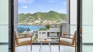 A balcony with two wooden chairs and a small table, overlooking a marina with boats and a mountainous town in the background.