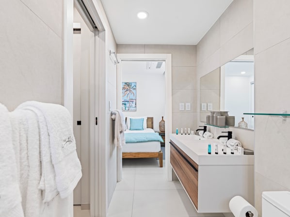 A modern bathroom with a large mirror, a white sink, and a toilet. There is a bed with a blue pillow and a painting of palm trees on the wall. - The Hills Residence (Simpson Bay)