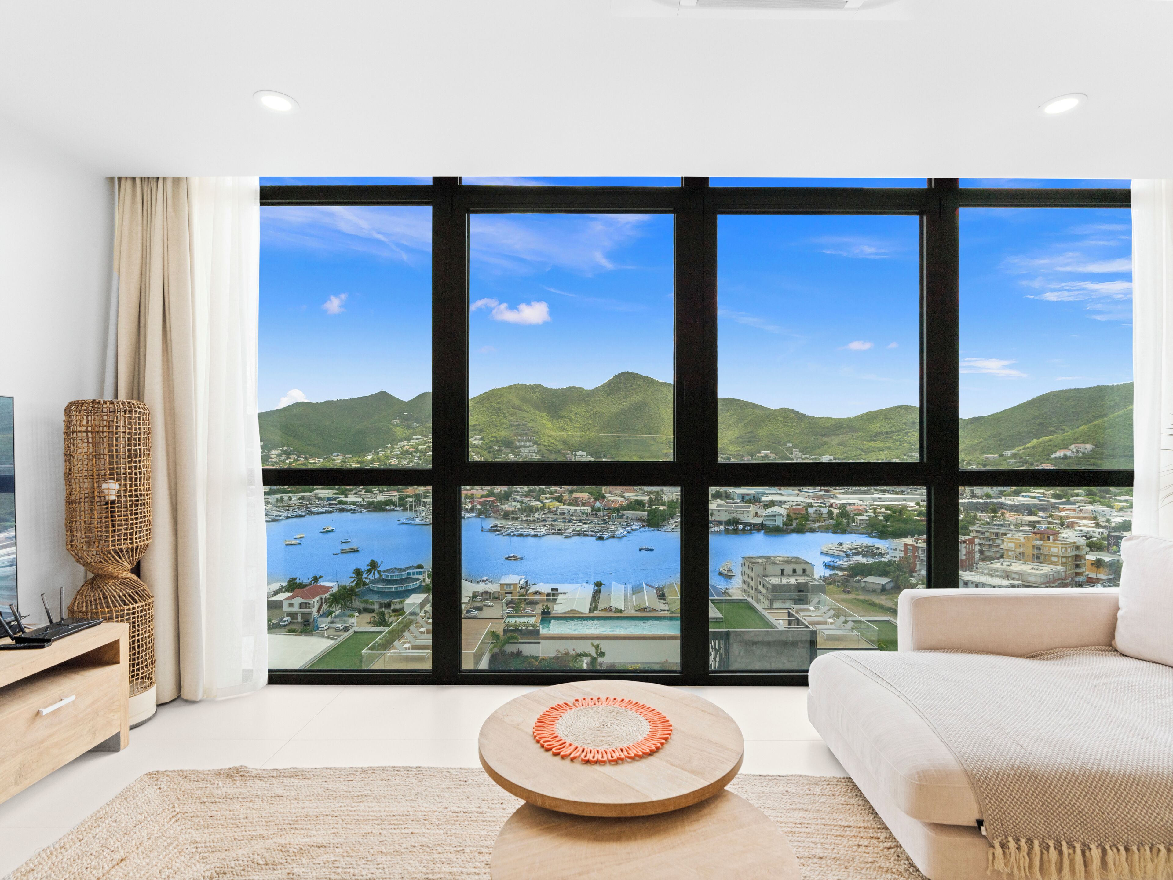 A modern living room with a large window offering a view of a coastal town and mountains.