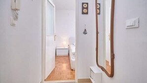 Kapea käytävä, jossa on puulattia, valkoinen ovi, joka johtaa makuuhuoneeseen, ja peili oikeanpuoleisella seinällä.
