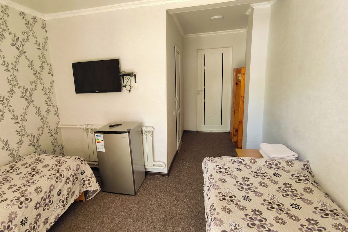 A small, simple room with two single beds, a small fridge, a TV mounted on the wall, and a door leading to another room.