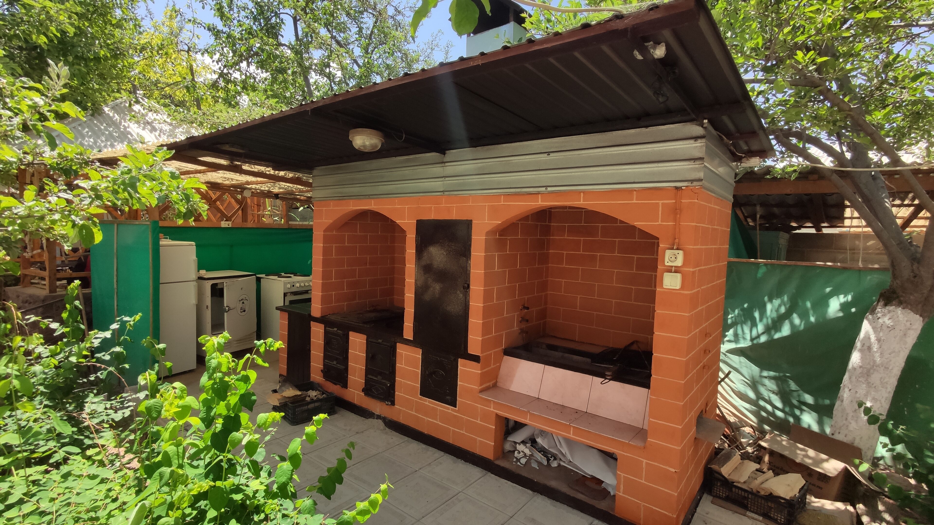 A brick-built outdoor kitchen with a metal roof, a green wall, and a wooden fence.
