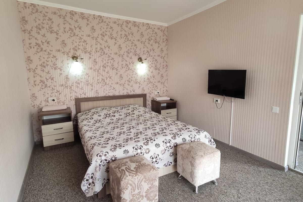 A bedroom with a bed, two bedside tables, a flat-screen TV, and a sliding glass door leading to a balcony.