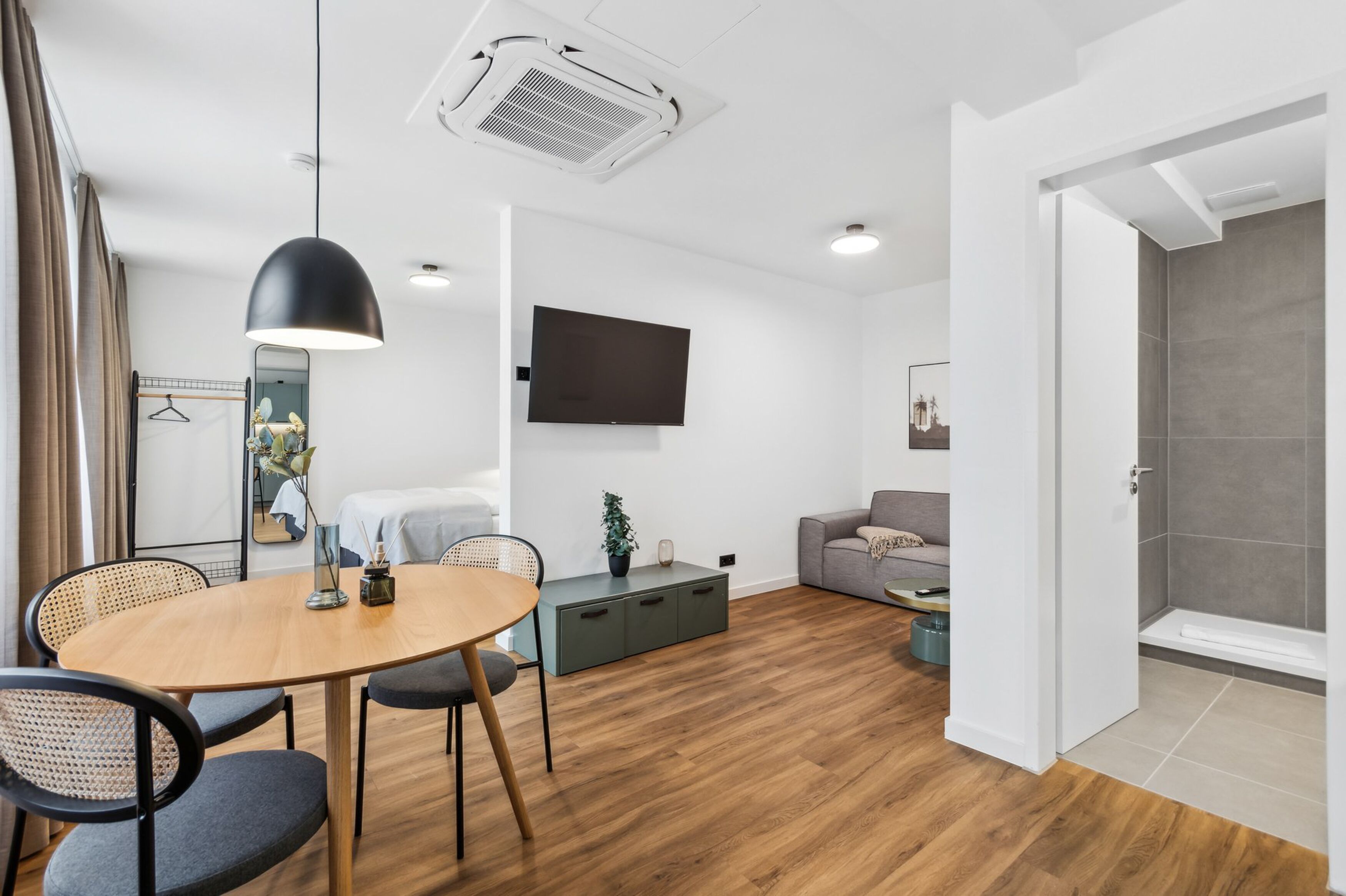 A modern hotel room with a wooden floor, a round dining table with chairs, a sofa, a TV mounted on the wall, and a shower area visible through an open door.