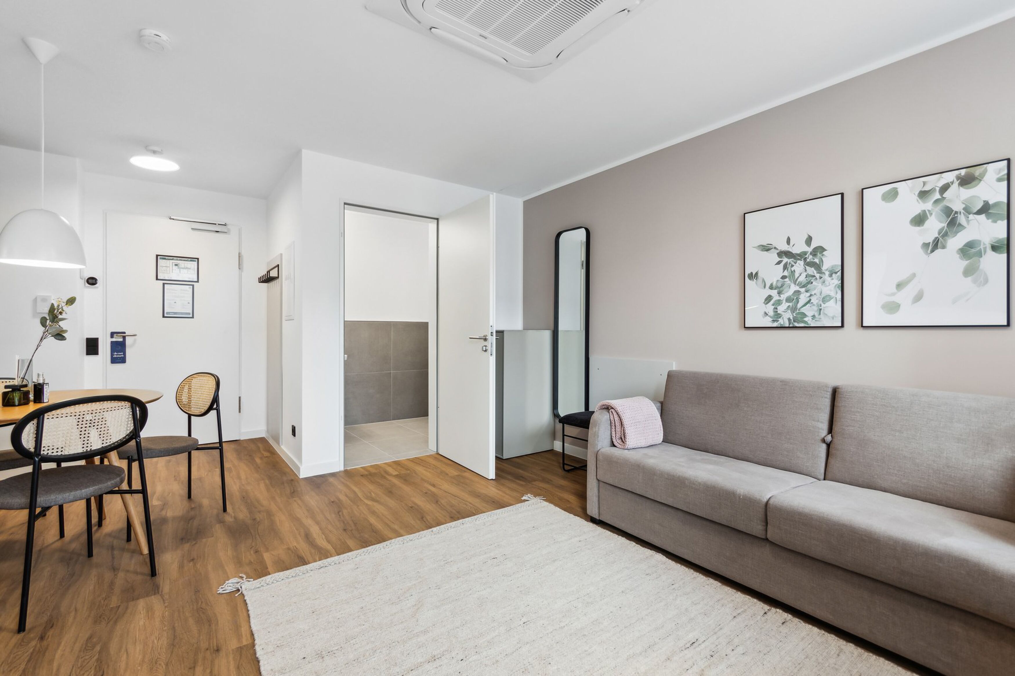 Ein modernes Wohnzimmer mit einem grauen Sofa, einem Esstisch mit Stühlen, einem weißen Teppich und zwei gerahmten Pflanzenbildern an der Wand.