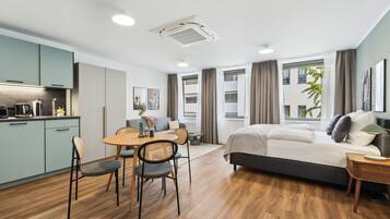Ein modernes Hotelzimmer mit einem Bett, einer Essecke mit Tisch und Stühlen, einer Küchenzeile und großen Fenstern mit Vorhängen.