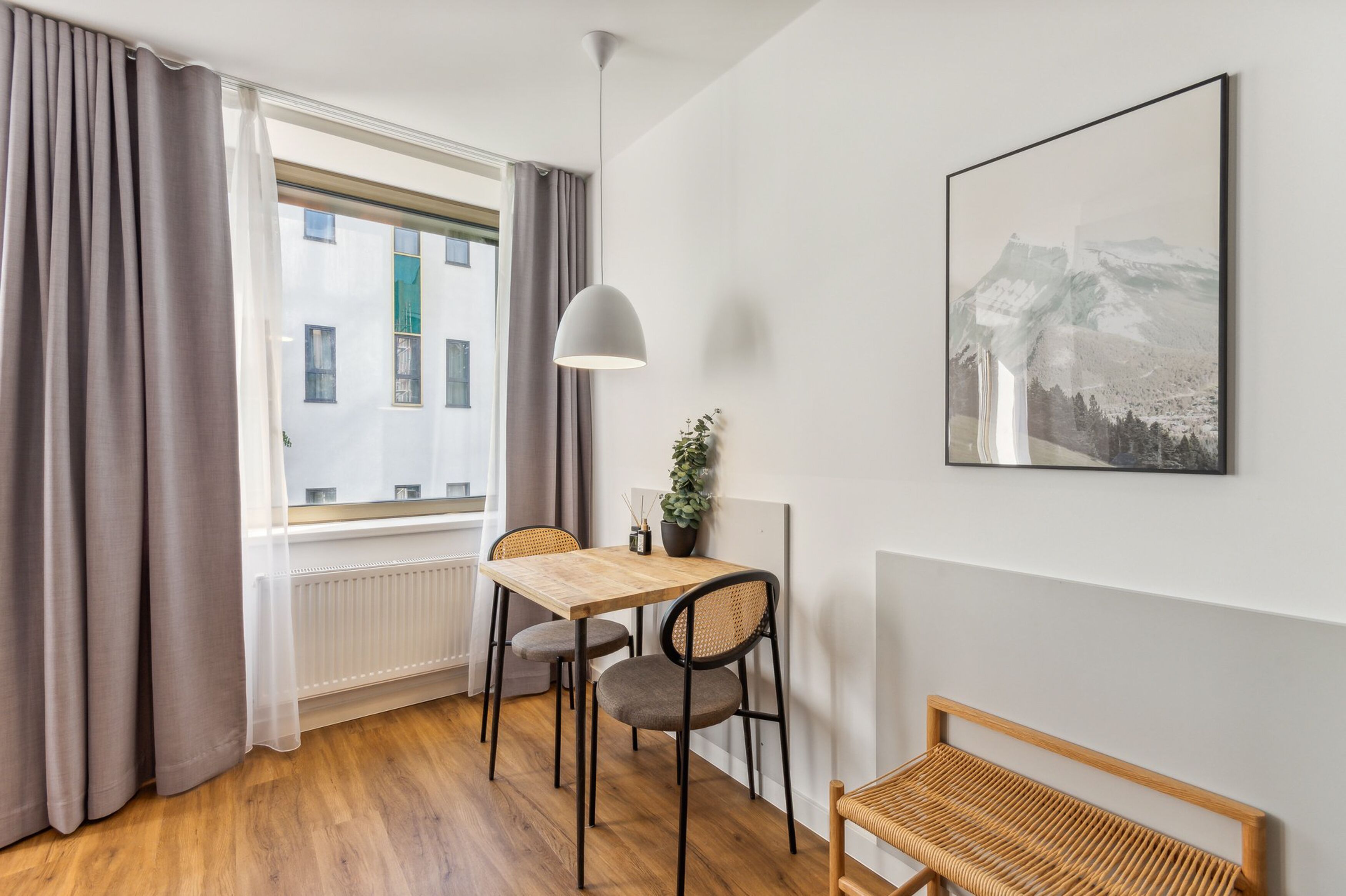 Um quarto com uma mesa de madeira, duas cadeiras, um banco, uma luminária pendente, uma planta em vaso e um quadro na parede.