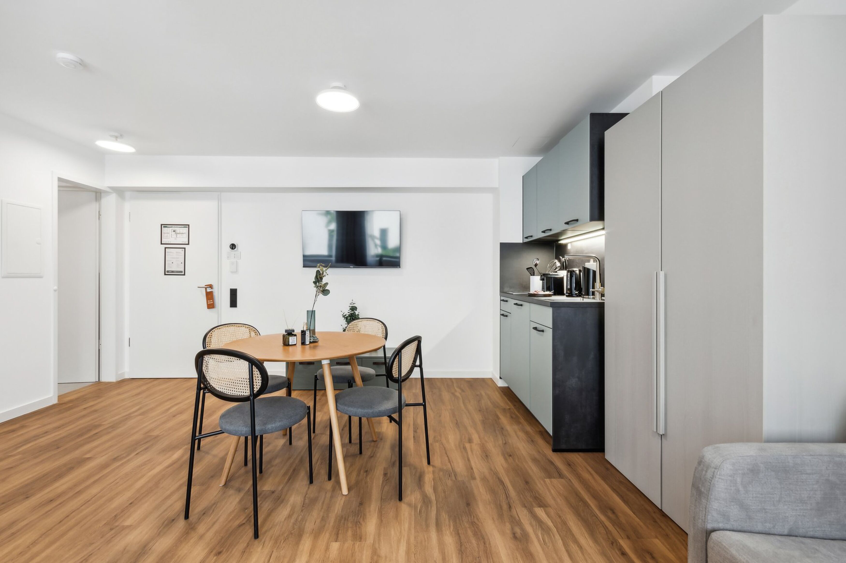 A modern kitchen with a dining area, featuring a wooden table, chairs, and a flat-screen TV mounted on the wall.
