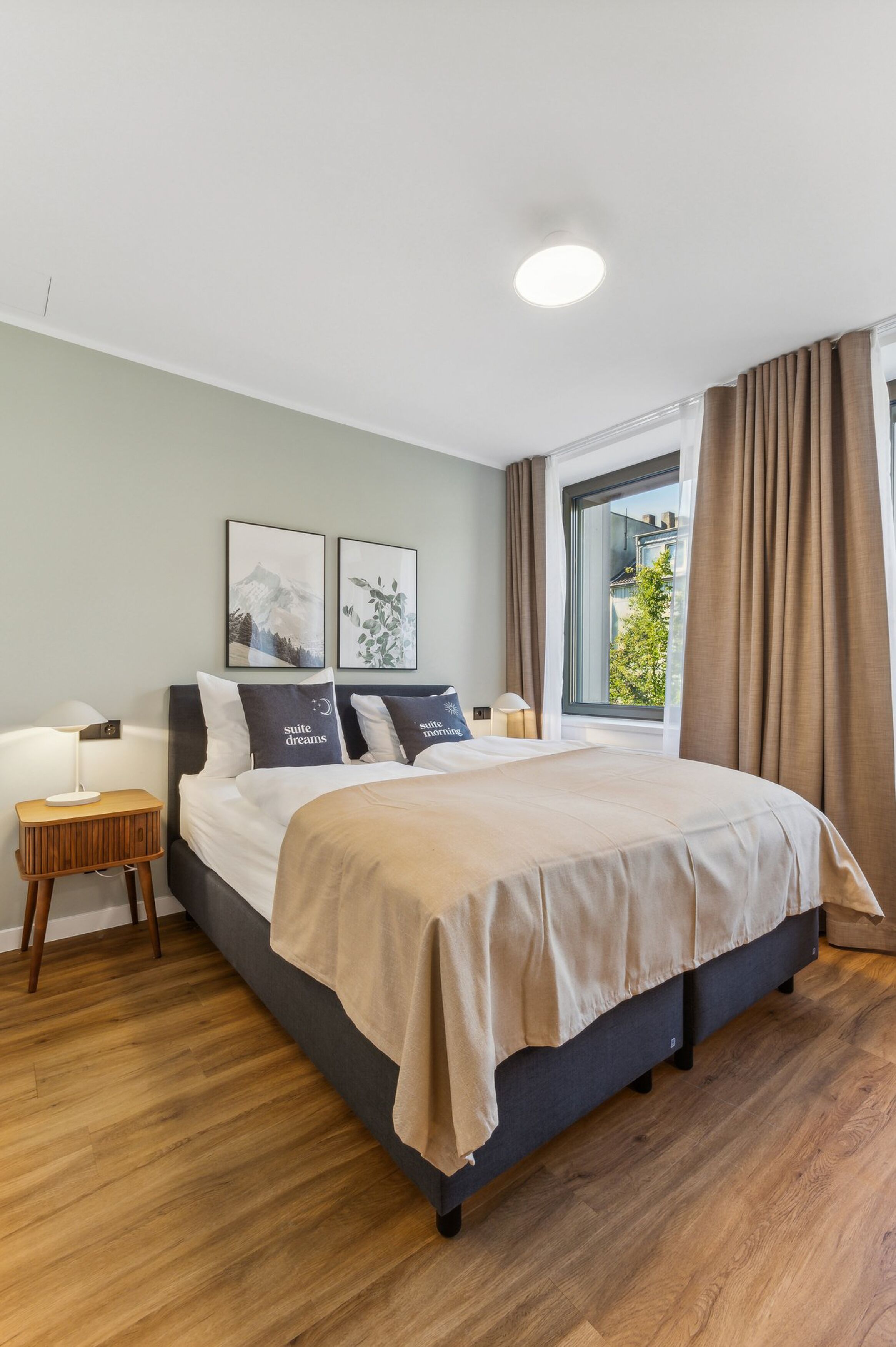 Una habitación de hotel moderna con una cama grande, suelo de madera, una ventana con cortinas y dos mesitas de noche con lámparas.