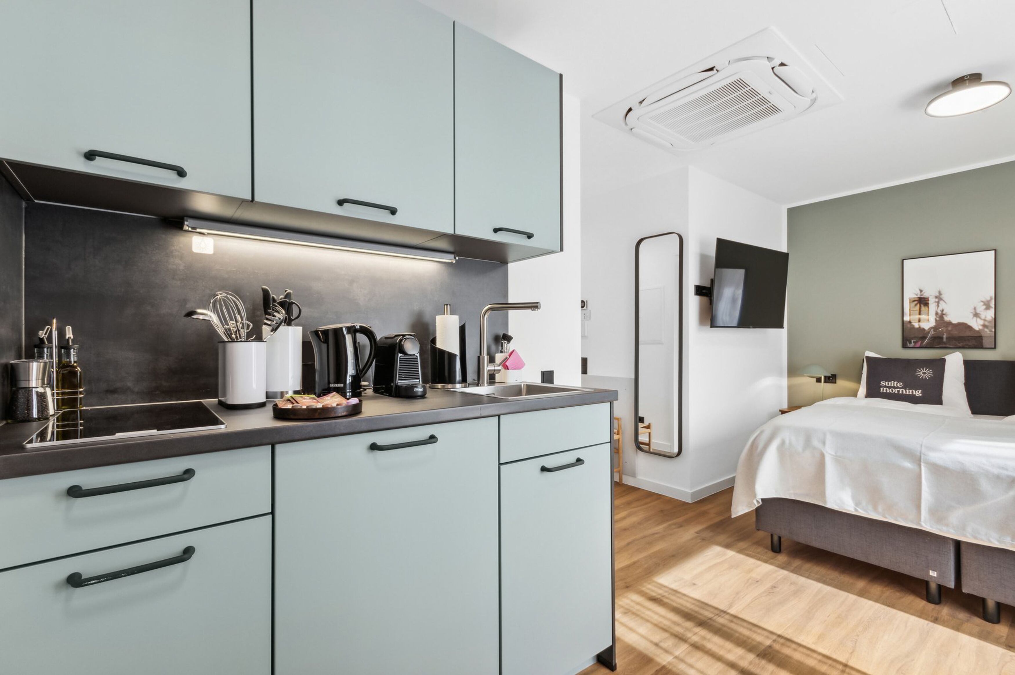 A modern kitchen with light blue cabinets, a black countertop, and various appliances. There is a bed with a white comforter and a pillow with "suite morning" text.