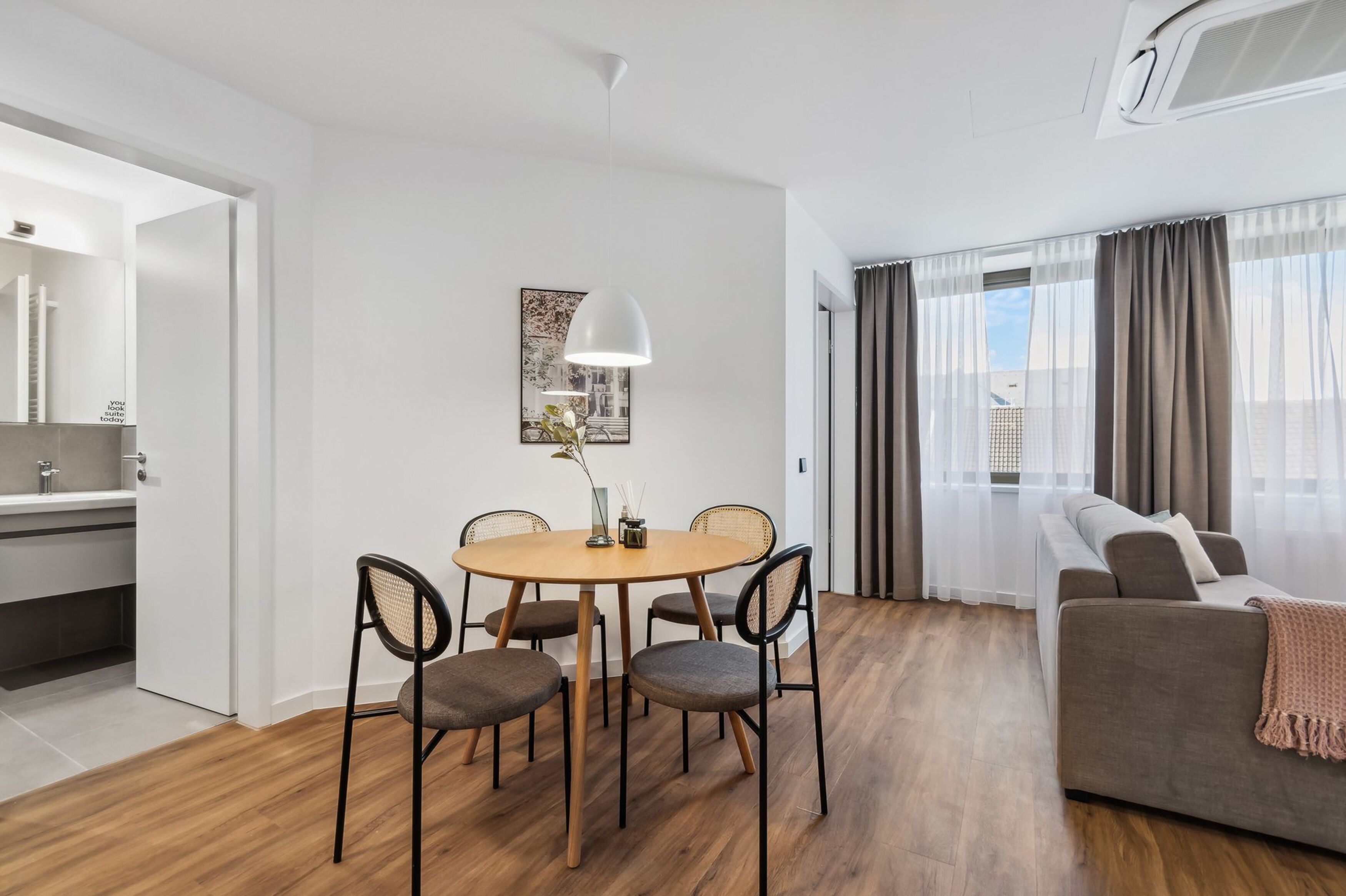 Een moderne woonkamer met een ronde houten tafel, vier stoelen, een bank en een groot raam met gordijnen.