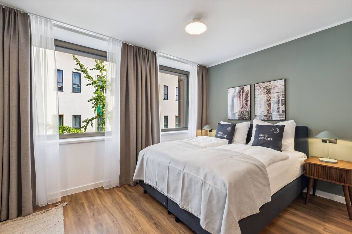 A hotel room with a large bed, two bedside tables, a window with curtains, and a view of buildings outside.