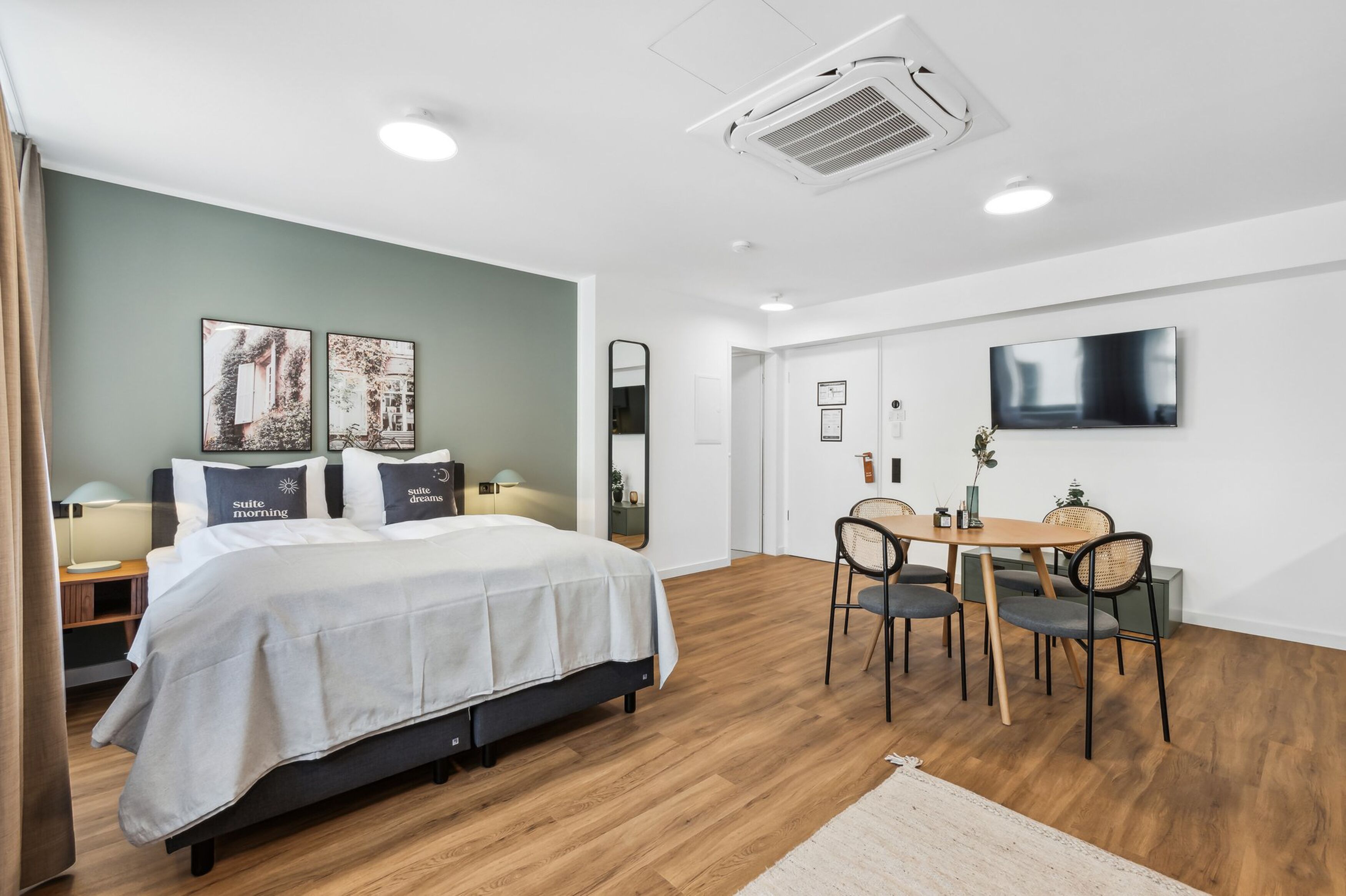 A modern hotel room with a bed, bedside table, lamp, and a dining area with chairs and a table.