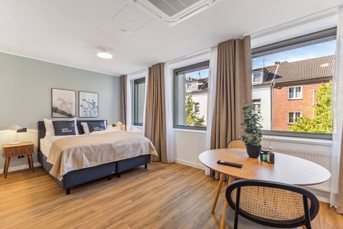 A hotel room with a bed, bedside table, lamp, and a view of a building through large windows.