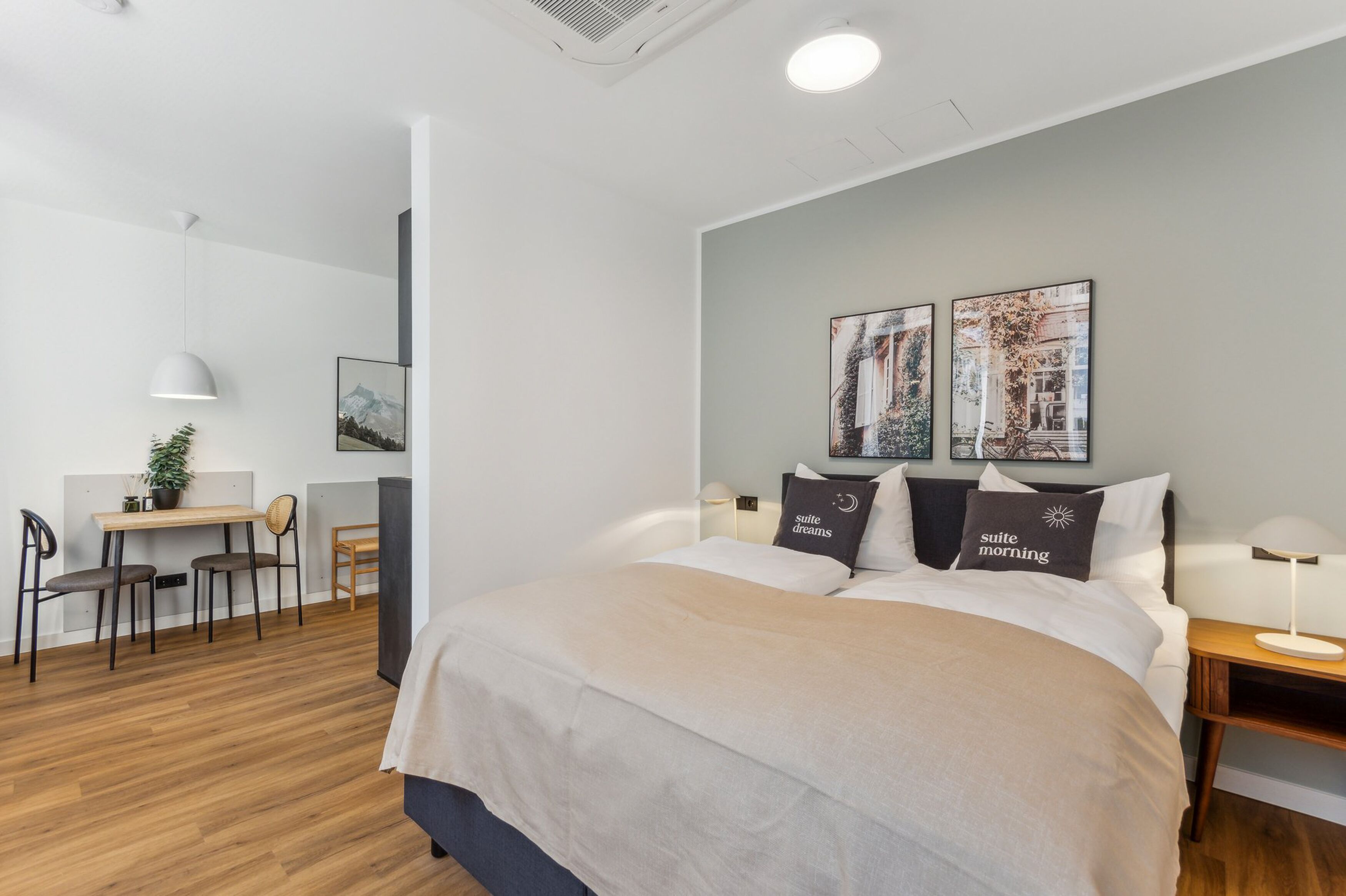A modern hotel room with a large bed, a desk, and two chairs. There are two wall-mounted artworks and a small table with a plant.