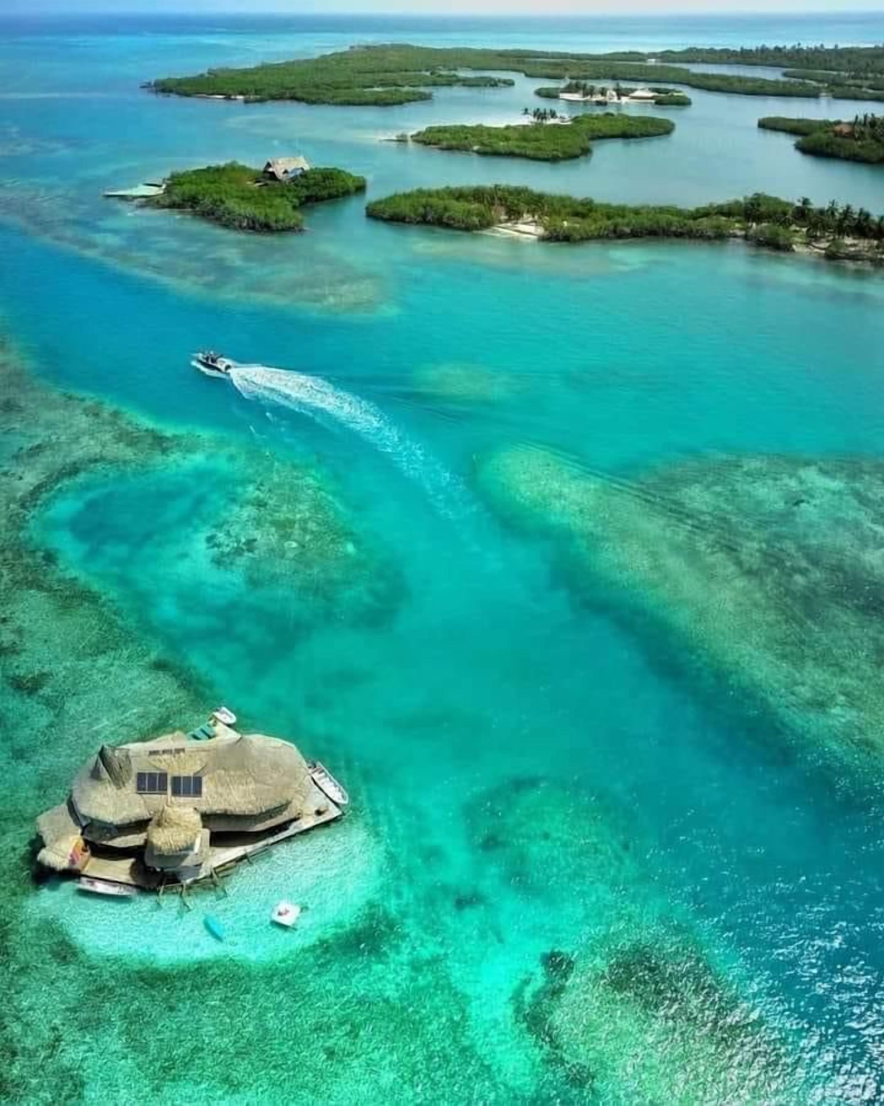 An aerial view of a tropical island with clear turquoise waters, a thatched-roof structure, and several smaller islands in the background.