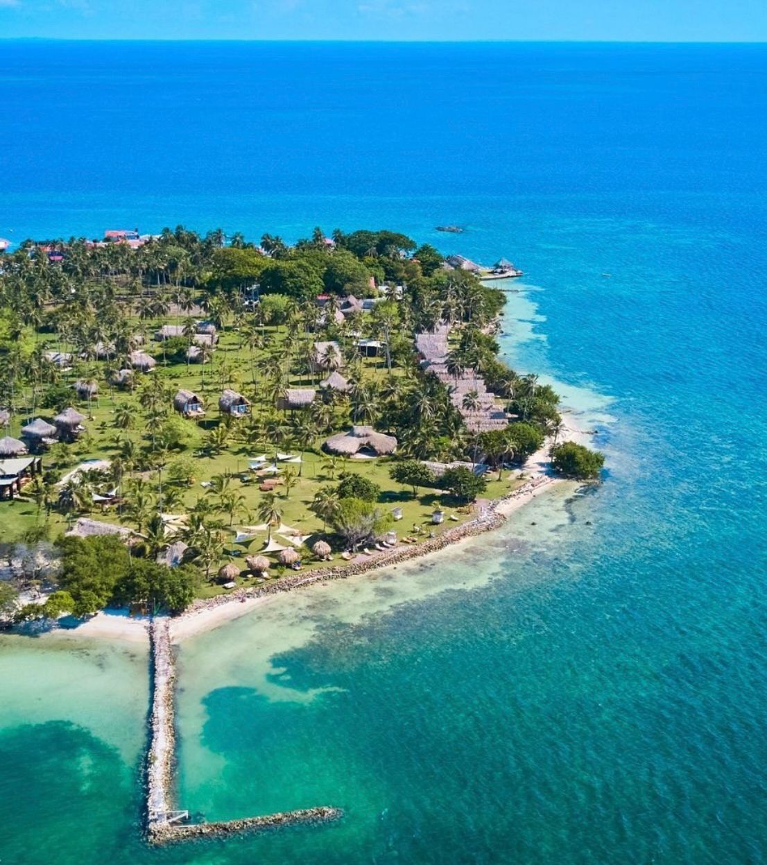 An aerial view of a tropical island with thatched-roof structures, clear turquoise waters, and a narrow beach.