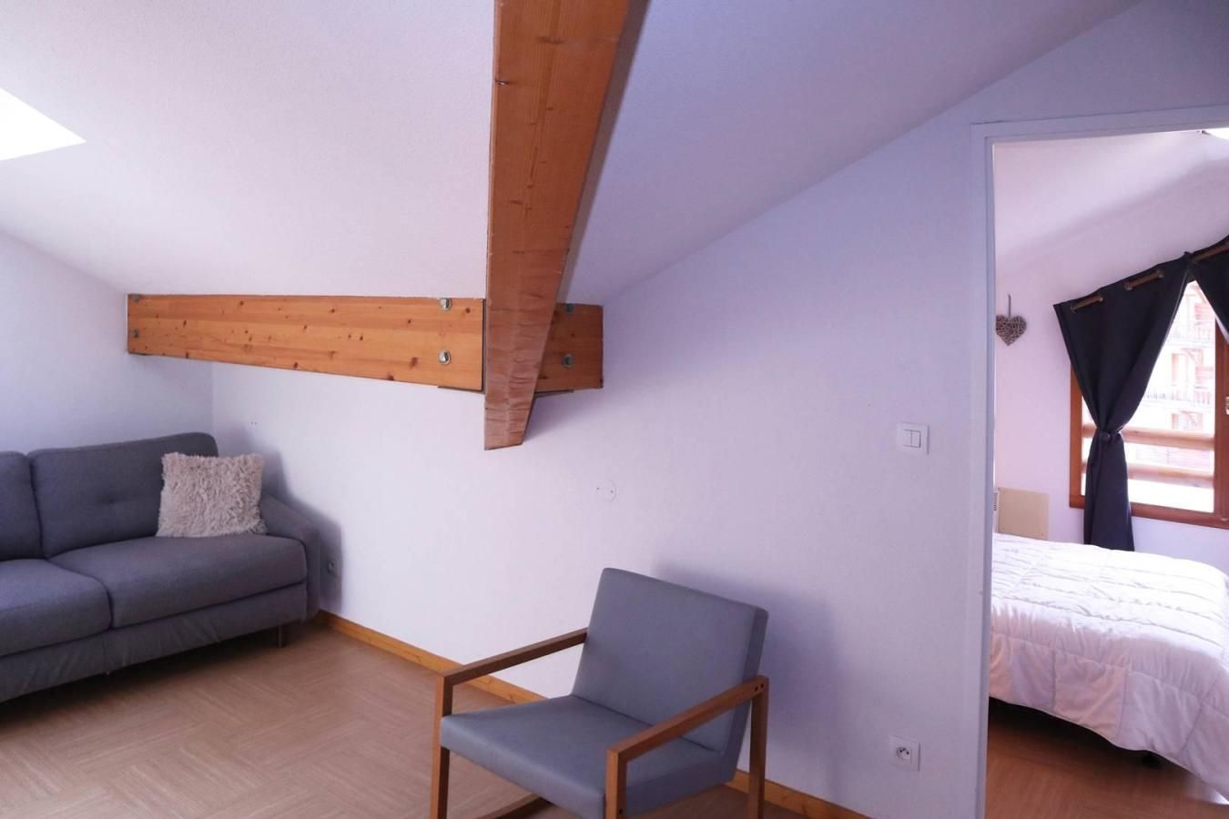 A room with a grey sofa, a wooden beam ceiling, a grey chair, and a bed with white bedding.
