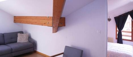 A room with a grey sofa, a wooden beam ceiling, a grey chair, and a bed with white bedding.