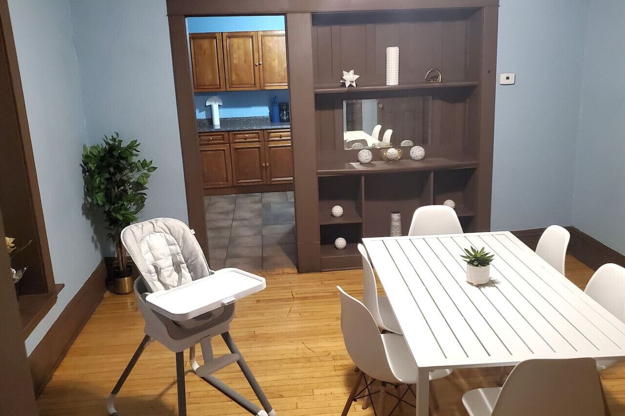 A dining area with a white table and chairs, a high chair, a potted plant, and a wooden cabinet with shelves.