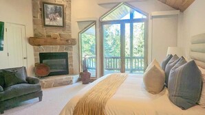 Un dormitorio con chimenea de piedra, una cama grande con muchas almohadas, un sillón oscuro y vistas al exterior a través de una puerta de vidrio.