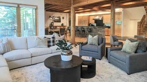 Amplia sala de estar con un sofá grande, dos sillones y una mesa de centro. Se ve una cocina y comedor al fondo.