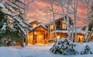 Une cabane en rondins avec une façade en pierre, entourée d’arbres couverts de neige et sous un ciel dégagé.