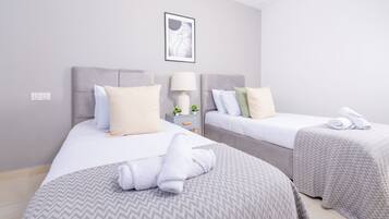 Une chambre d’hôtel avec deux lits, chacun doté d’une tête de lit grise, de draps blancs et d’une couvre-lit grise à motifs. Une table de chevet avec une lampe et une petite plante se trouve entre les deux lits.