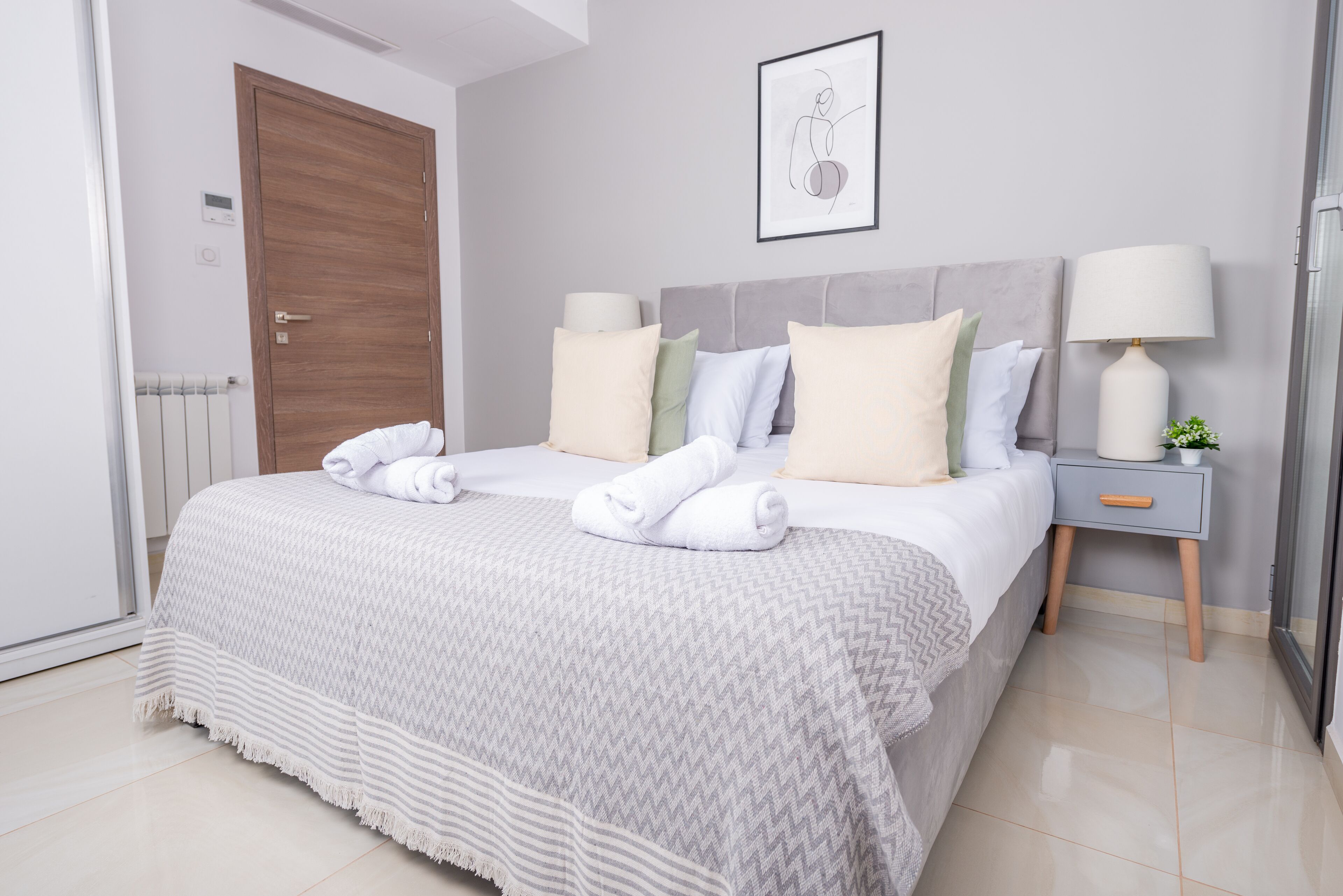 A neatly made bed with a grey and white patterned bedspread, two bedside tables with lamps, and a framed artwork on the wall.