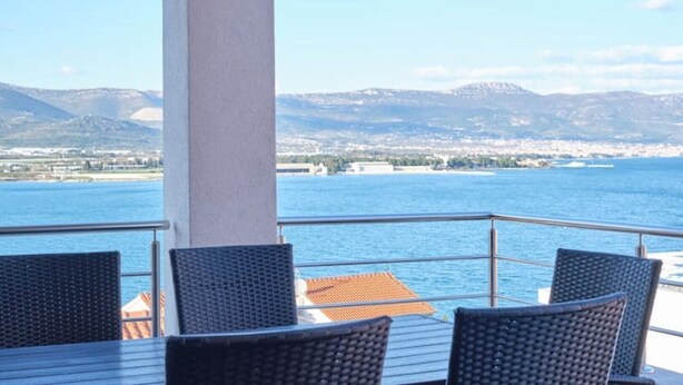 A balcony with wicker chairs and a table, overlooking a body of water and a distant cityscape. - Queen's Lodge Mastrinka by Litto (Mastrinka)