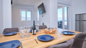 A dining area with a wooden table set for four, a television mounted on the wall, and a view of the sea through large windows. - Queen's Lodge Mastrinka by Litto (Mastrinka)