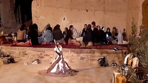 A group of people gathered around a fire in a rustic outdoor setting with stone walls and a brick floor.