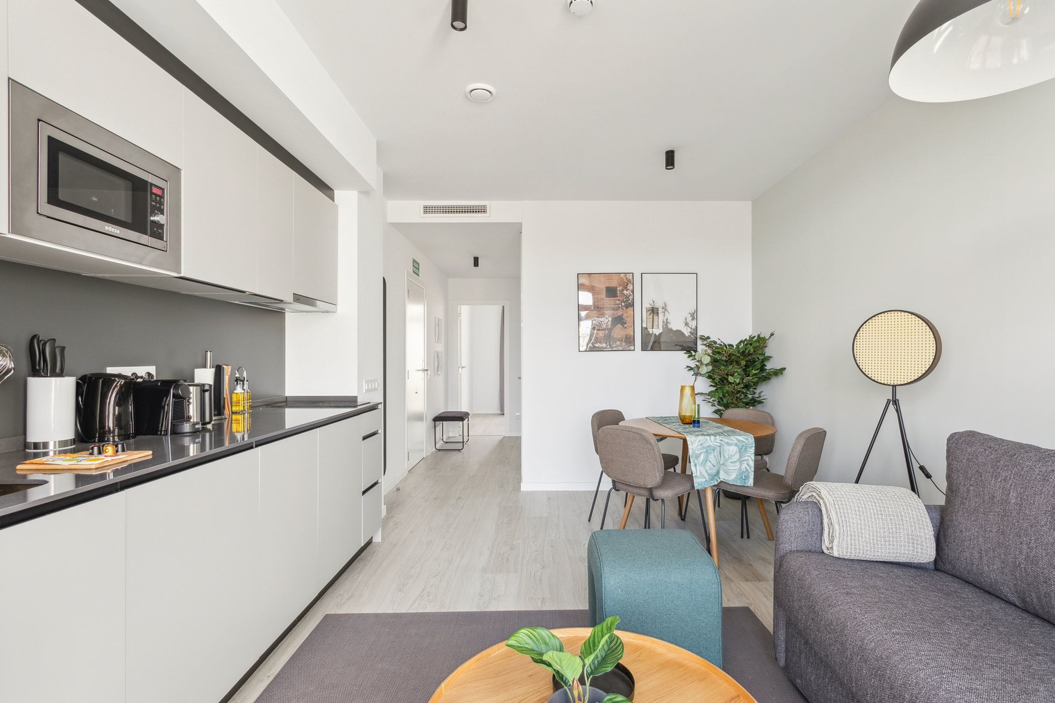 A modern kitchen with a microwave, coffee maker, and kettle. There is a dining area with a table and chairs, and a sofa.