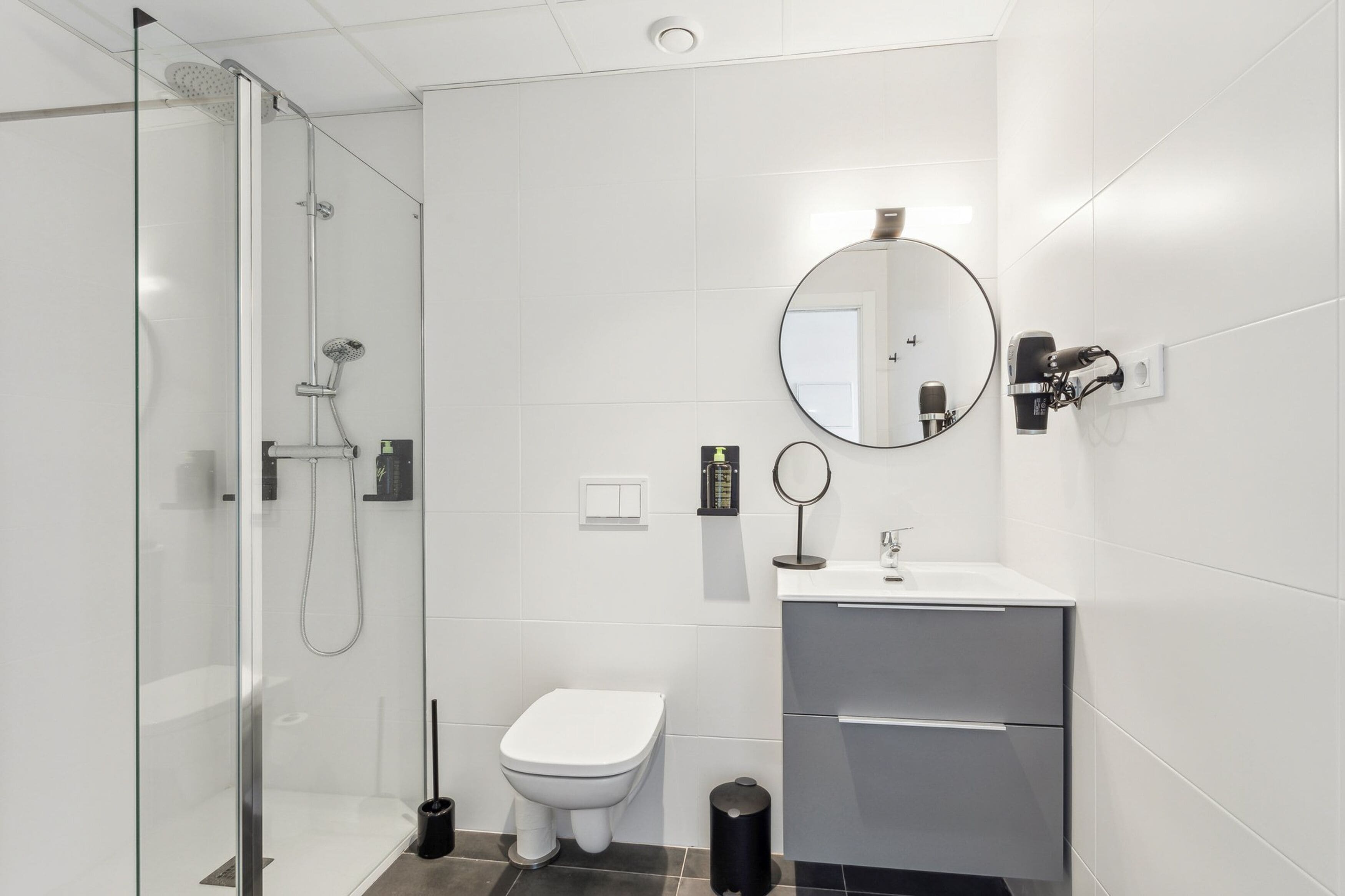 Un baño moderno con ducha envuelta en vidrio, inodoro blanco, lavamanos con espejo y un mueble pequeño.